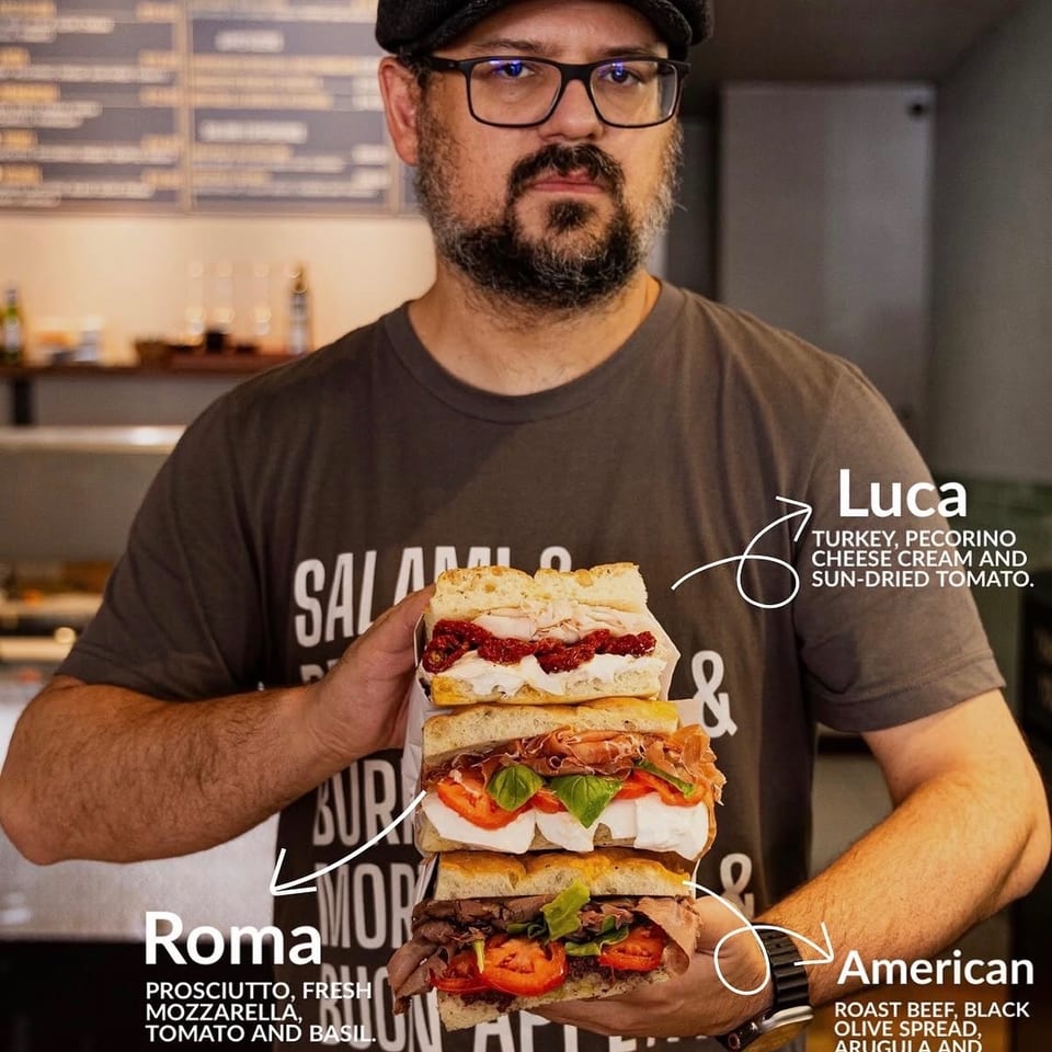 Man in a flat cap and glasses holding a stack of three gourmet sandwiches from Turci Panino.