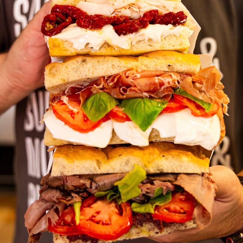 A person holding a towering stack of three focaccia sandwiches with various fillings.