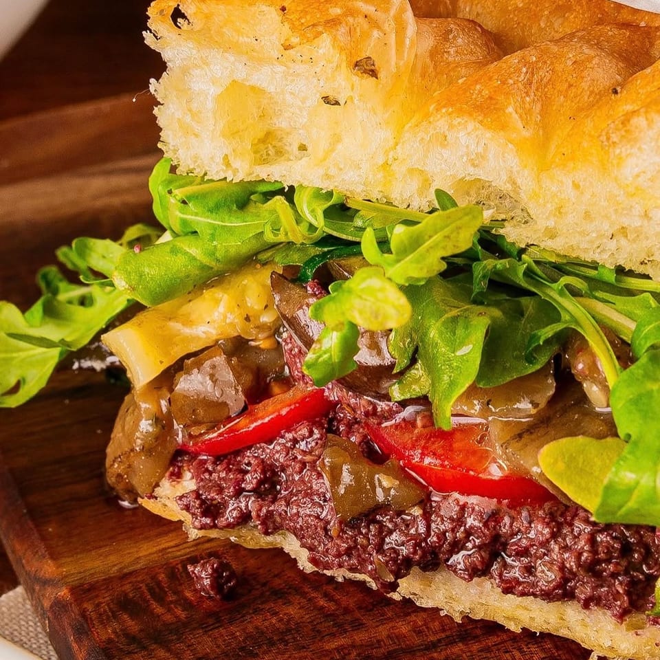 Close-up of a focaccia sandwich filled with olive tapenade, roasted peppers, and arugula on a wooden board.